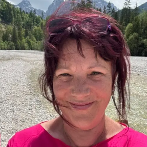 Eine lächelnde Frau mit dunklen Haaren und pinkfarbenem Oberteil steht vor einer alpinen Flusslandschaft mit Bergen im Hintergrund.
